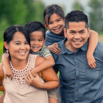 Photo of a smiling family. The parents are each carrying a child on their back.