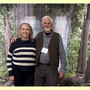 Our executive director, Bev, stands and smiles with Kurt Hoelting, our event speaker. They stand in front of a cloth backdrop with a photo of a cedar forest and nurse log.