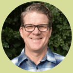 David smiles in front of a leafy tree. He wears a blue plaid button-down shirt and dark framed glasses. He has short, light brown, wavy hair combed back.