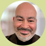 Rich smiles at the camera. He is bald with a short salt and pepper beard and moustache. He wears a dark brown blazer over a black t-shirt.