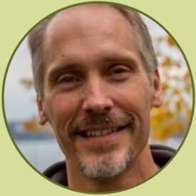 Scott stands in front of an autumn tree and a blurry body of water. He has a short goatee and short blonde and silver hair. He wears a brown sweater.