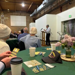 Guests at the 2025 Spring Luncheon sit around round tables draped in Forest green table cloths. Lilacs blossom out of small glass vases on each table. Former Board President Rob Caudillo addresses the guests. Behind him is a backdrop of a cedar forest with a nurse log.