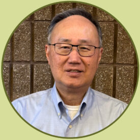 Vic stands in front of a brick wall and smiles at the camera. He wears glasses with rectangular frames and a light blue button-up shirt.
