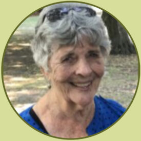 Peggy smiles at the camera. She is seated by the trunk of a tree. She has short, silver hair and laugh lines dancing across her face. She wears a blue blouse and sunglasses tucked on top of her head.