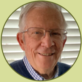 Doug smiles at the camera. He has short, white hair and wears glasses, a sweater, and a button-up shirt with a grid pattern on it.