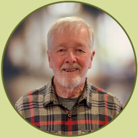 Larry smiles at the camera. He is wearing a grey and red plaid flannel shirt. He has short white hair and a short white beard.