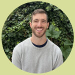 Andrew stands in front of a wall of green ivy and smiles brightly. He has short, brown hair and a short beard. He wears a grey sweater.