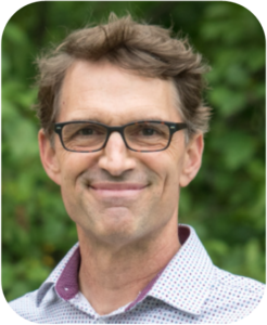 Photo portrait of Carl HiltonVanOsdall. Carl smiles in front of green foliage. He has short, light brown hair. He wears glasses and a button-down shirt.