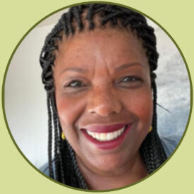 Deb smiles at the camera. She has a rich, coffee-toned complexion and long black box braids. She wears red lipstick and a grey blouse.