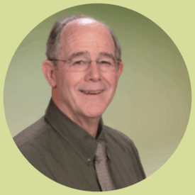 Gary smiles at the camera. He has short, grey hair, rectangular glasses without frames, and he wears an olive colored button-up shirt with a light brown tie.