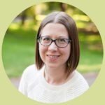 Grace sits in a grassy park, smiling at the camera. She has straight, brown, shoulder length hair and wears teal glasses. She is wearing a thick cream colored sweater.