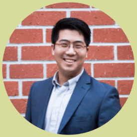 Isaiah stands in front of a red brick wall, smiling. He has short, black hair and wears rectangular glasses with red frames. He wears a navy blue suit and light blue button-up shirt.