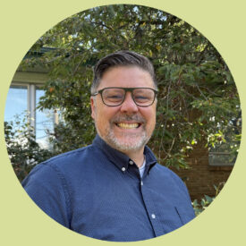 Jim smiles in front of the Ravenna office. He has short, grey-brown hair and a short salt and pepper beard. He wears a navy blue button up shirt and glasses.