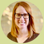 Katherine smiles at the camera under dappled sunlight. She has shoulder-length copper hair, freckles, and wears glasses and a modest brown v-neck sweater.