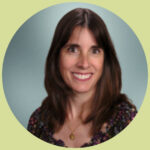 Tita smiles at the camera in front of a faded blue background. She has long, wavy brown hair and wears a patterned blouse.