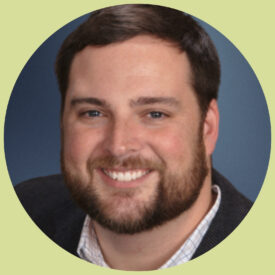 Matt smiles at the camera. He has short, brown hair and blue eyes. He has a short beard and wears a button-down shirt with a light grid pattern on it.