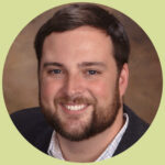 Matt smiles at the camera. He has short, brown hair, a short beard, and blue eyes. He wears a button-up shirt with a grid pattern and a dark grey blazer over.